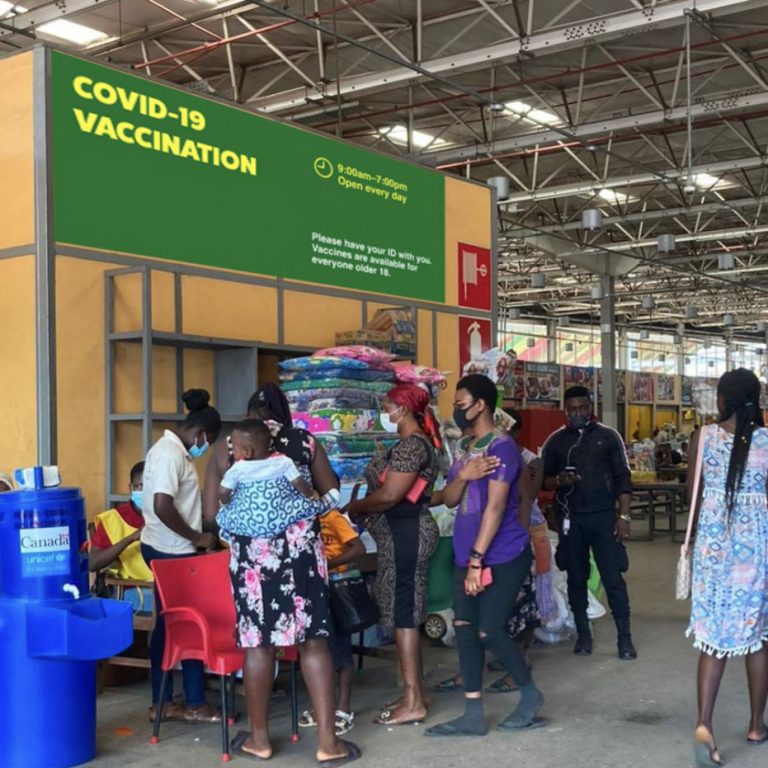 Mockup of a green and yellow banner hanging above the vaccination pop-up in Kumasi City Market that reads "COVID-19 Vaccination" in big text with opening hours and "Please have your ID with you. Vaccines are available to everyone over 18" in smaller text.