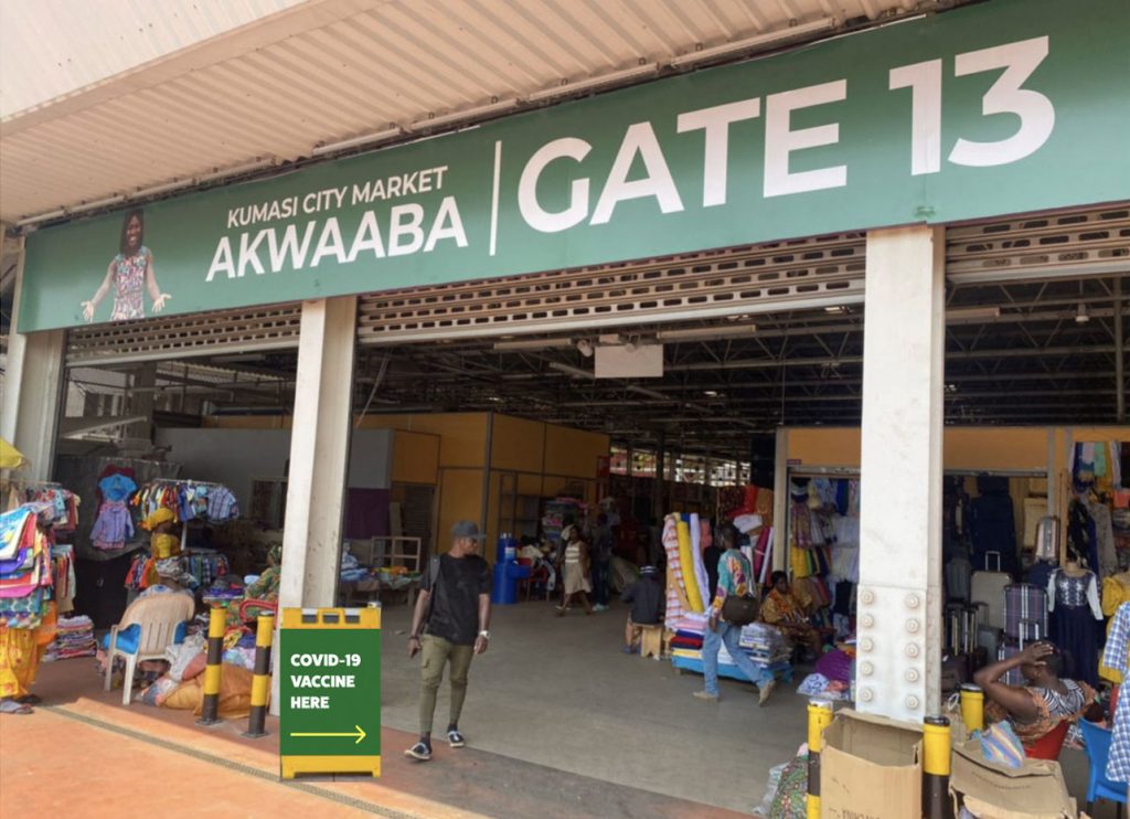 Mockup of a green and yellow sandwich board sign with text that reads "COVID-19 Vaccine Here" with an arrow pointing inside Kumasi City Market.